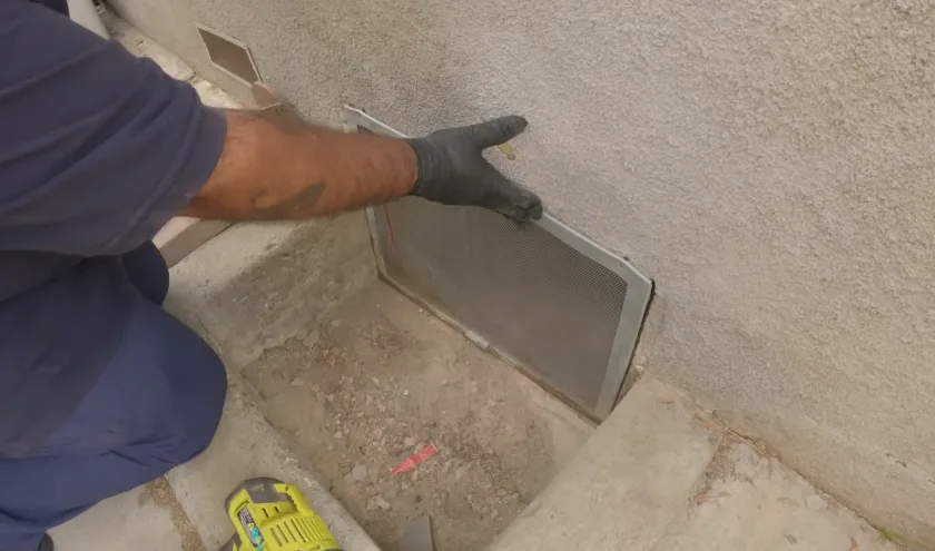 Gloved hand holding a metal vent cover screen before refitting it to a wall opening